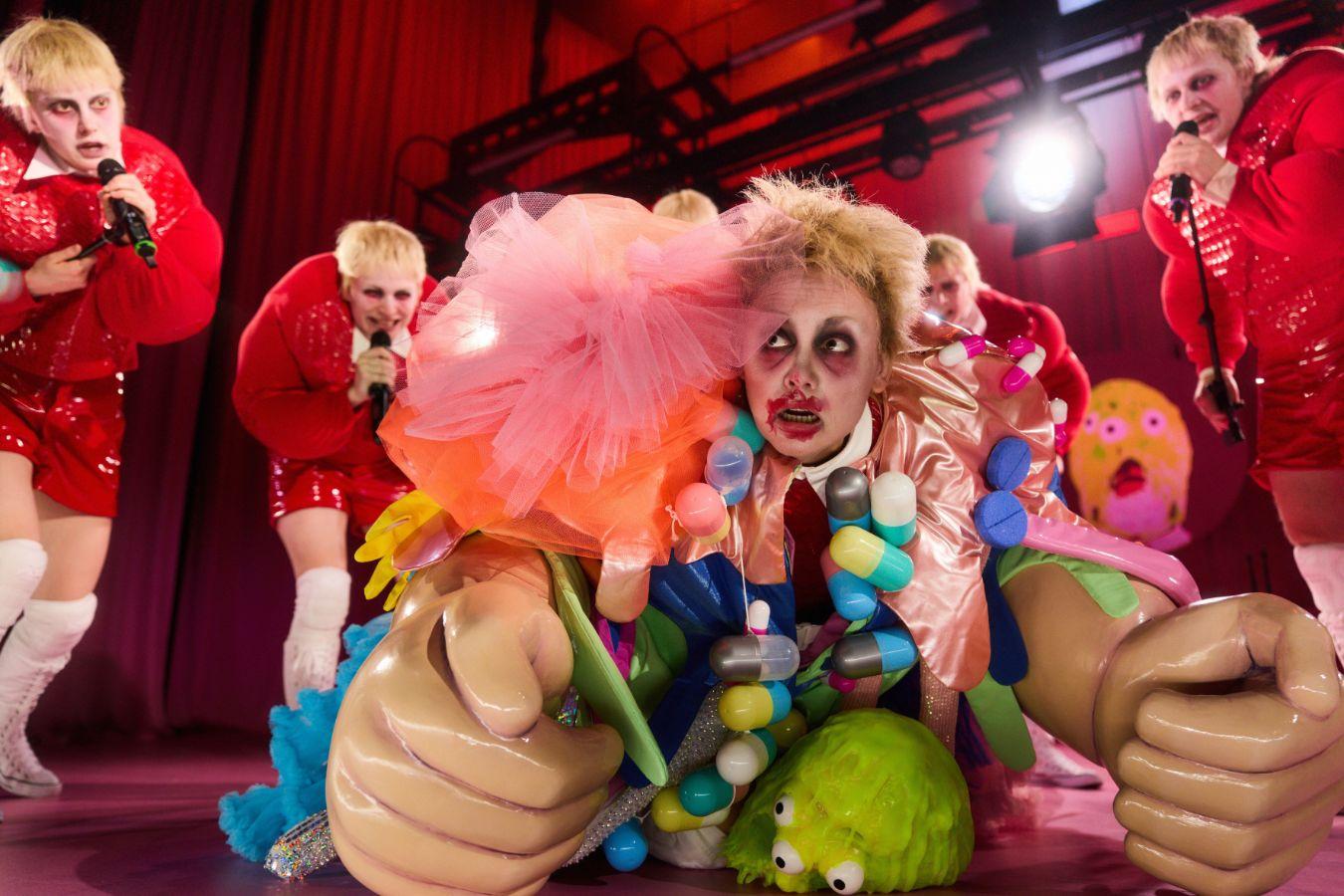 Bühnenperformance mit mehreren Personen in glitzernden roten Kostümen; im Vordergrund eine farbenfroh verkleidete Figur mit großen, überdimensionierten Requisitenhänden und buntem Dekor, umgeben von grellem Bühnenlicht.