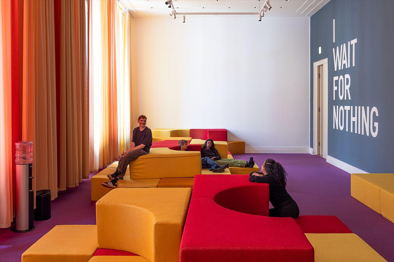 Eine Aufnahme von einem Raum mit lila Teppichboden, in dem sich verschiedene Polsterelemente in gelb und rot befinden, darauf sitzend vier Personen. Über die Länge der linken Wand ziehen sich gelbe und rote Vorhänge, die rechte Wand ist petrolfarben mit eine großen Schriftzug in weiß "I WAIT FOR NOTHING".