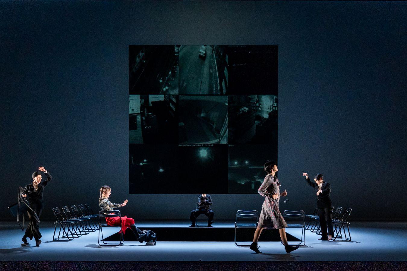 square opening in the stage floor, with a large screen behind it; rows of chairs around the opening and people moving through the space.