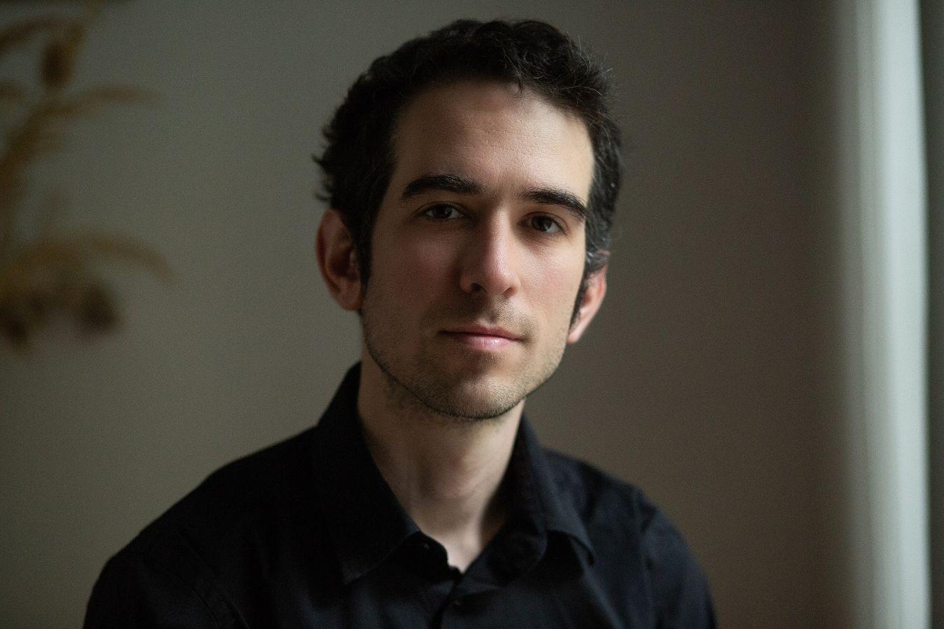 A man with short black hair and a black shirt looks kindly into the camera.