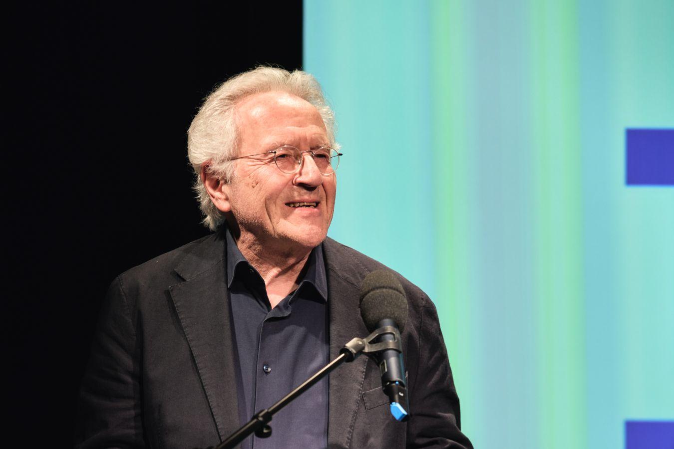 The dramaturg Hermann Beil has white hair and thin-rimmed glasses. He is wearing a dark shirt and jacket and is standing on a stage at the microphone.