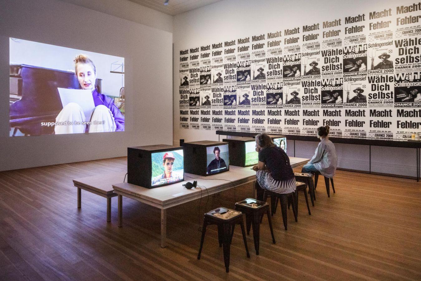 Personen sitzen vor Fernsehern in einer Ausstellung mit wandfüllenden Textplakaten.