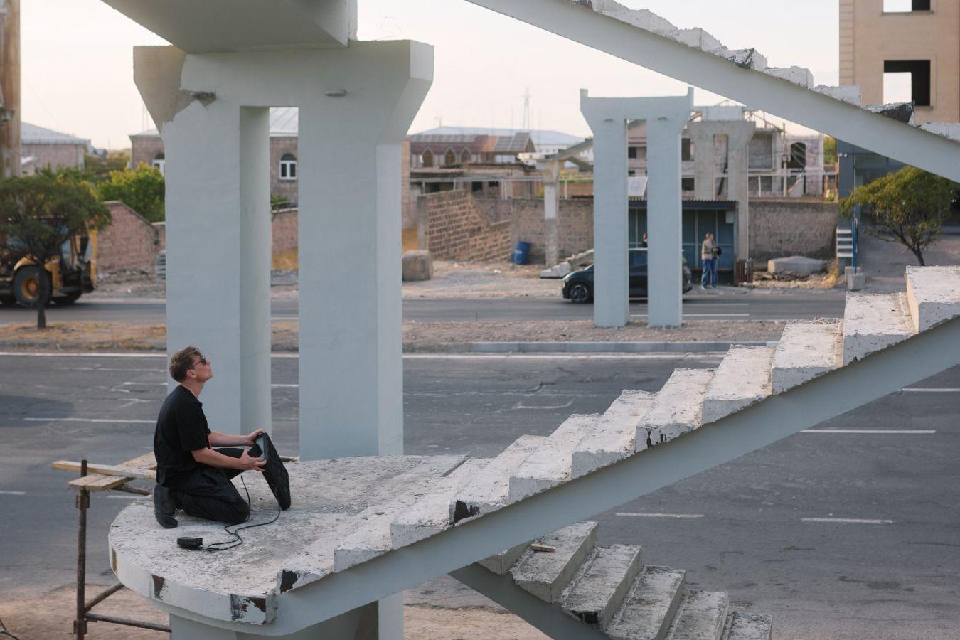 A person dressed in black sits with an electronic music device in the white concrete ruins of an unfinished bridge.