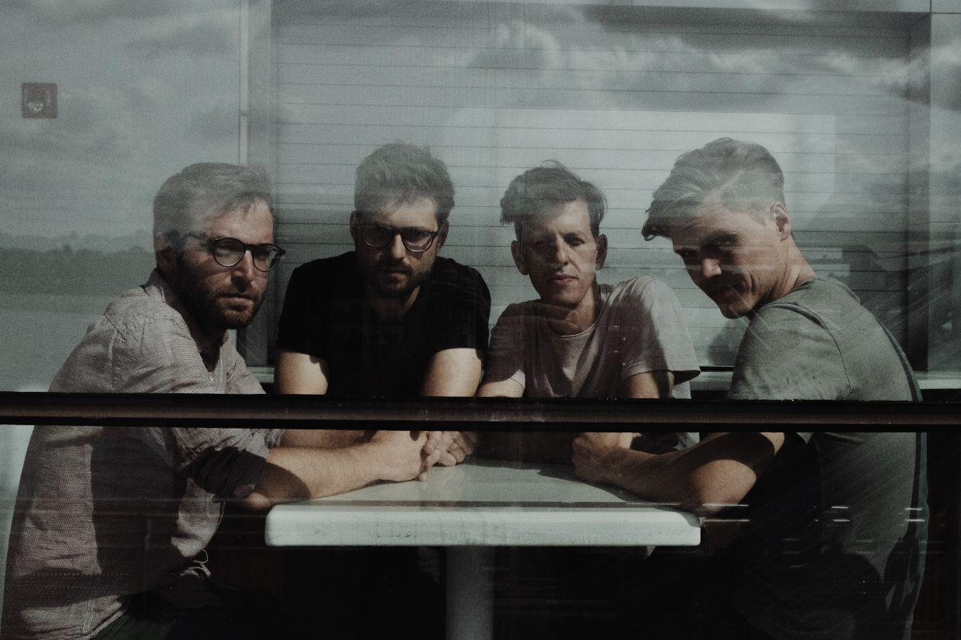 Die vier Musik*innen von Nikel sitzen an einem Tisch. Vor ihnen befindet sich eine Scheibe, die die Ansicht verzerrt.