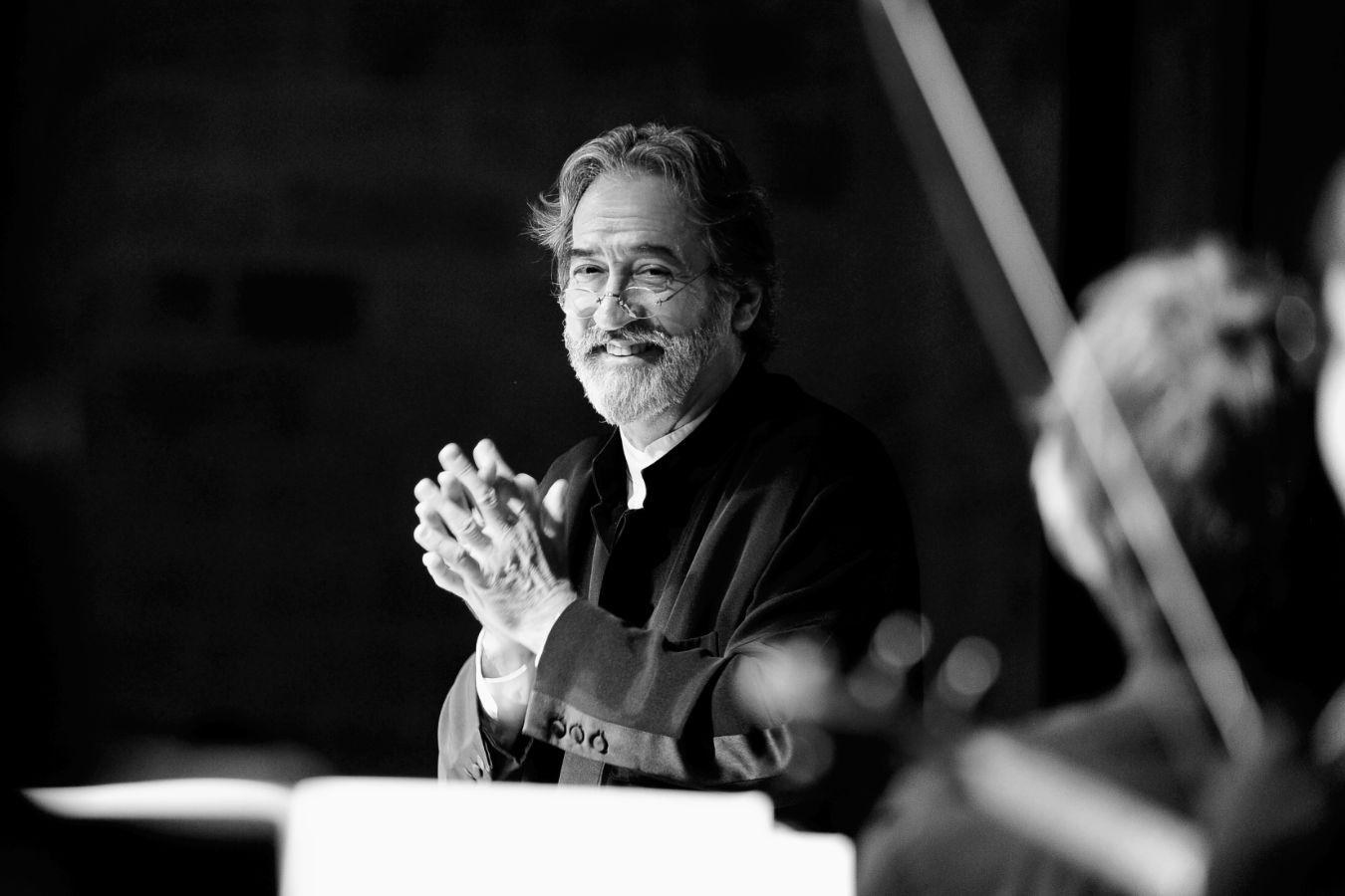 Ein älterer Mann mit Bart hält seine Hände vor der Brust verschränkt und lacht in die Kamera. Er steht vor einem Dirigentenpult an der Seite sind unscharf Streichinstrumente zu erkennen.