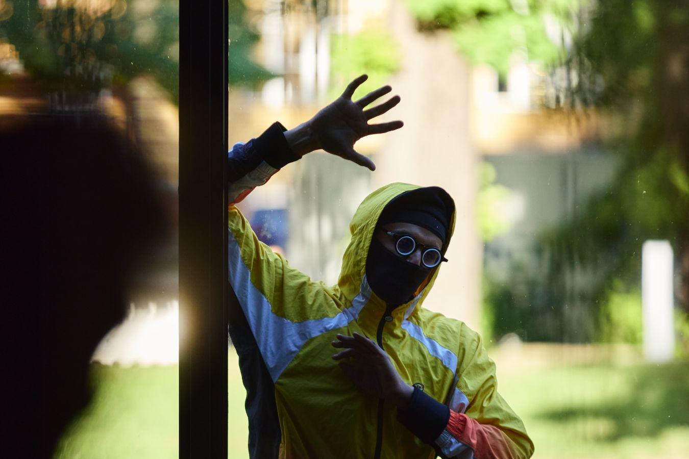 Behind a wall of windows, Kieron Jina can be seen with his arm raised at an angle and their hand spread open. They are wearing the hood of a green-yellow tracksuit top with light blue stripes and blue glasses. White vertical bars of varying lengths on the image indicate that an audio clip is available.