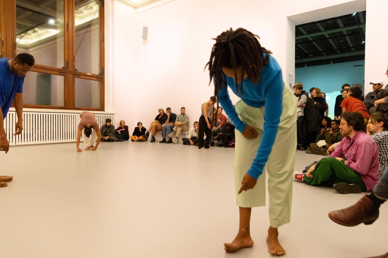 Several people perform in a light pink-painted room while an audience watches from benches, observing the dancers’ movements.