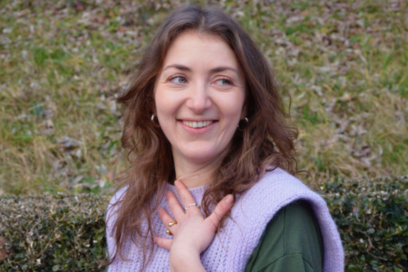 A woman with long brown hair in a purple knitted vest looking to the right and holding her hand to her chest