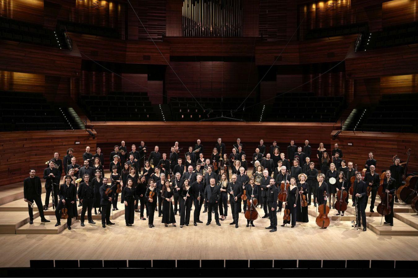 An orchestra stands with its instruments in a concert hall.