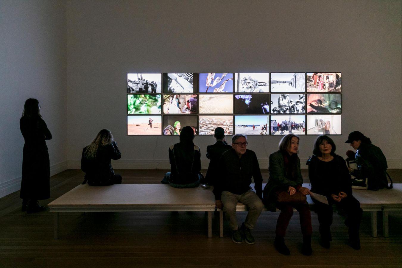 Personengruppe sitzt auf einer Bank vor einer Wand mit mehreren Bildschirmen, die verschiedene Bilder zeigen.