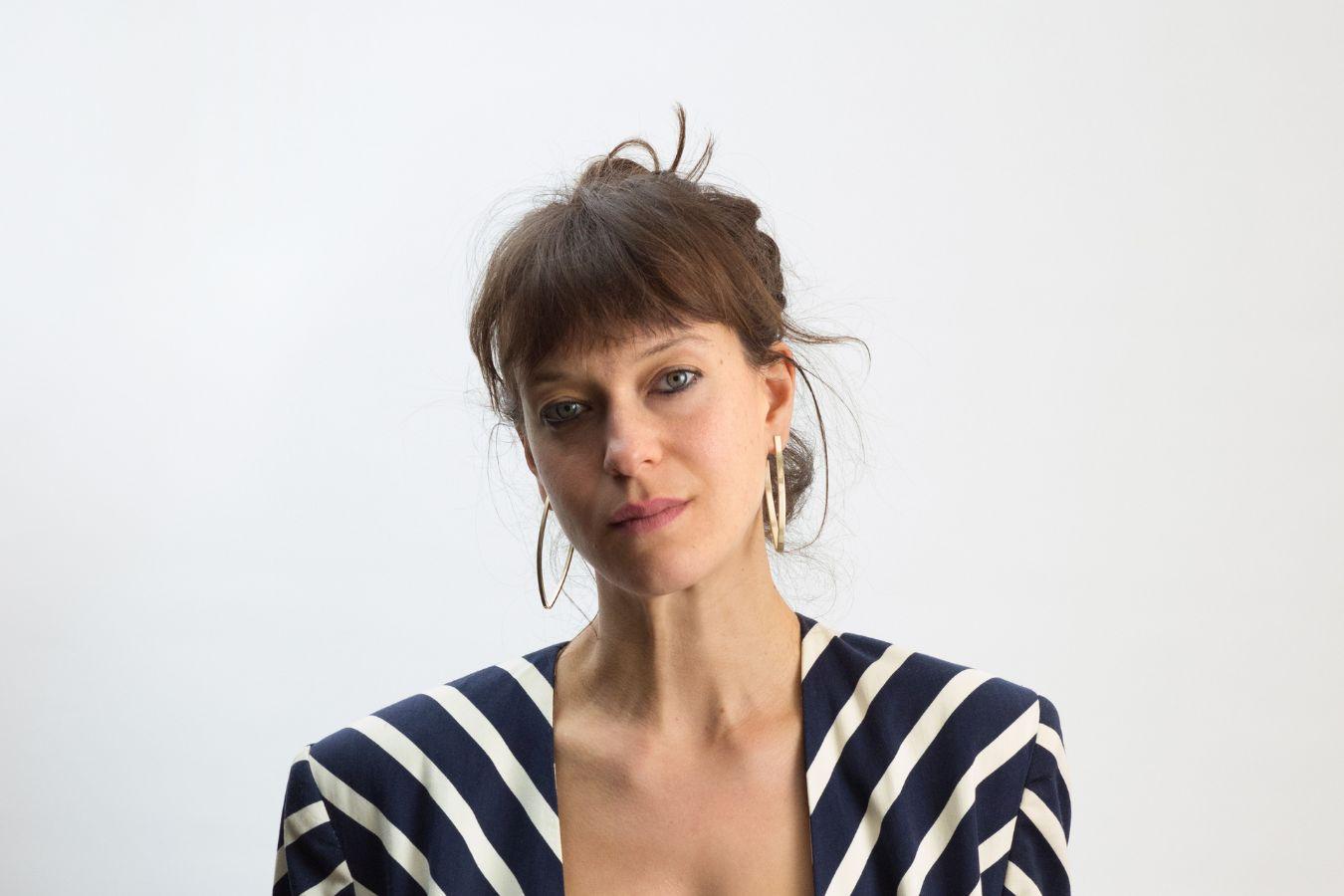 Halbporträt einer Person mit langen braunen Haaren im Zopf, trägt einen gestreiften Blazer.