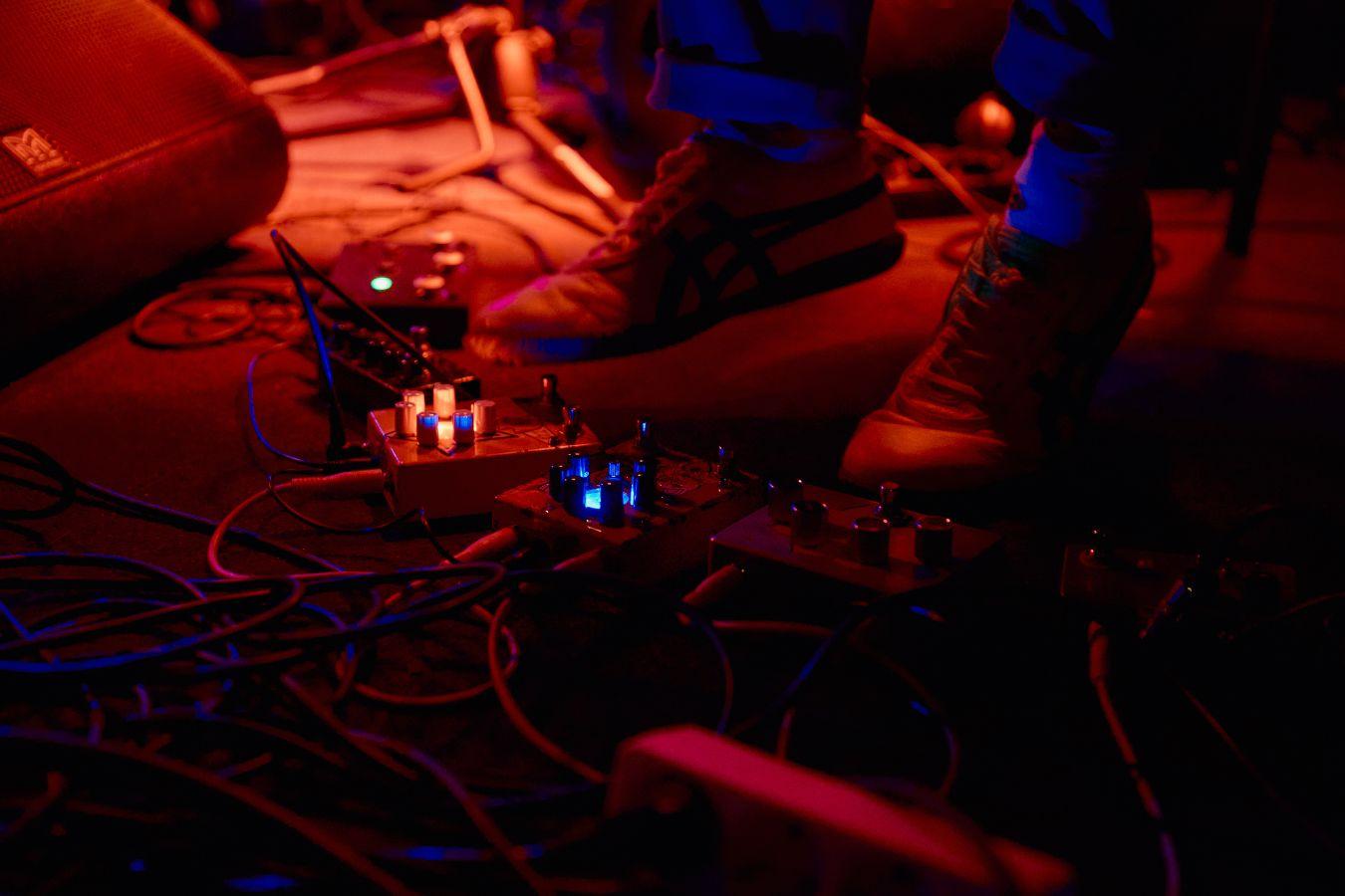 Feet among effect pedals on a stage in colorful light.