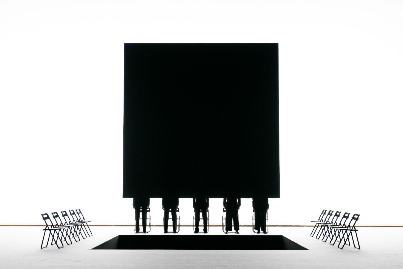 Eine weiße Bühne mit quadratischem Loch, dahinter eine große schwarze Leinwand, davor 5 schwarze Silouetten.