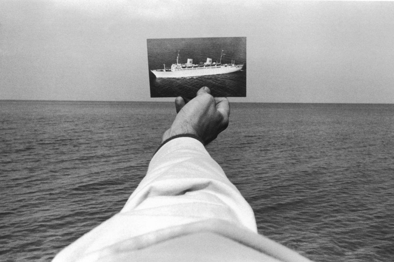 An outstretched arm holds a postcard showing a ship against the horizon of the sea.