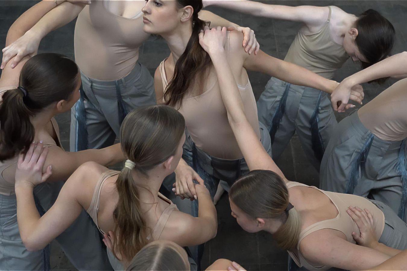 Several dancers stand close together in a room, their arms intertwined. They are wearing simple costumes with beige-colored tops.