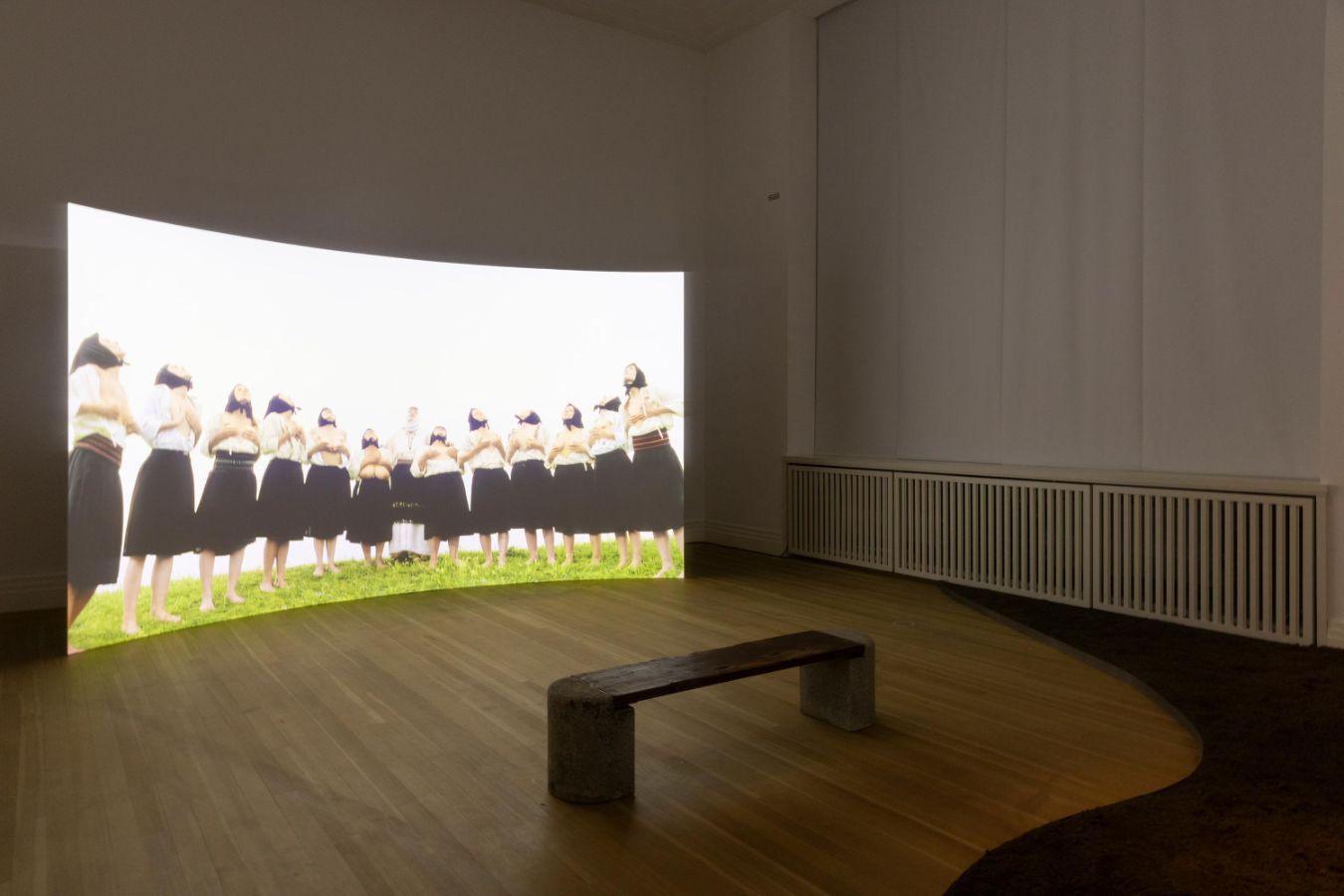 Im Ausstellungsraum steht eine Leinwand, auf der mehrere Personen stehen und gen Himmel schauen. Vor der Leinwand steht eine Bank.