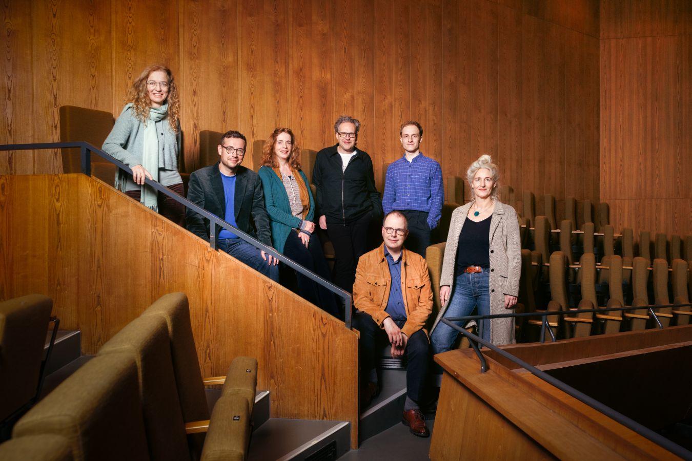 Die sieben Theatertreffen-Jurymitglieder stehen oder sitzen im Rang eines Theaters und schauen in die Kamera.