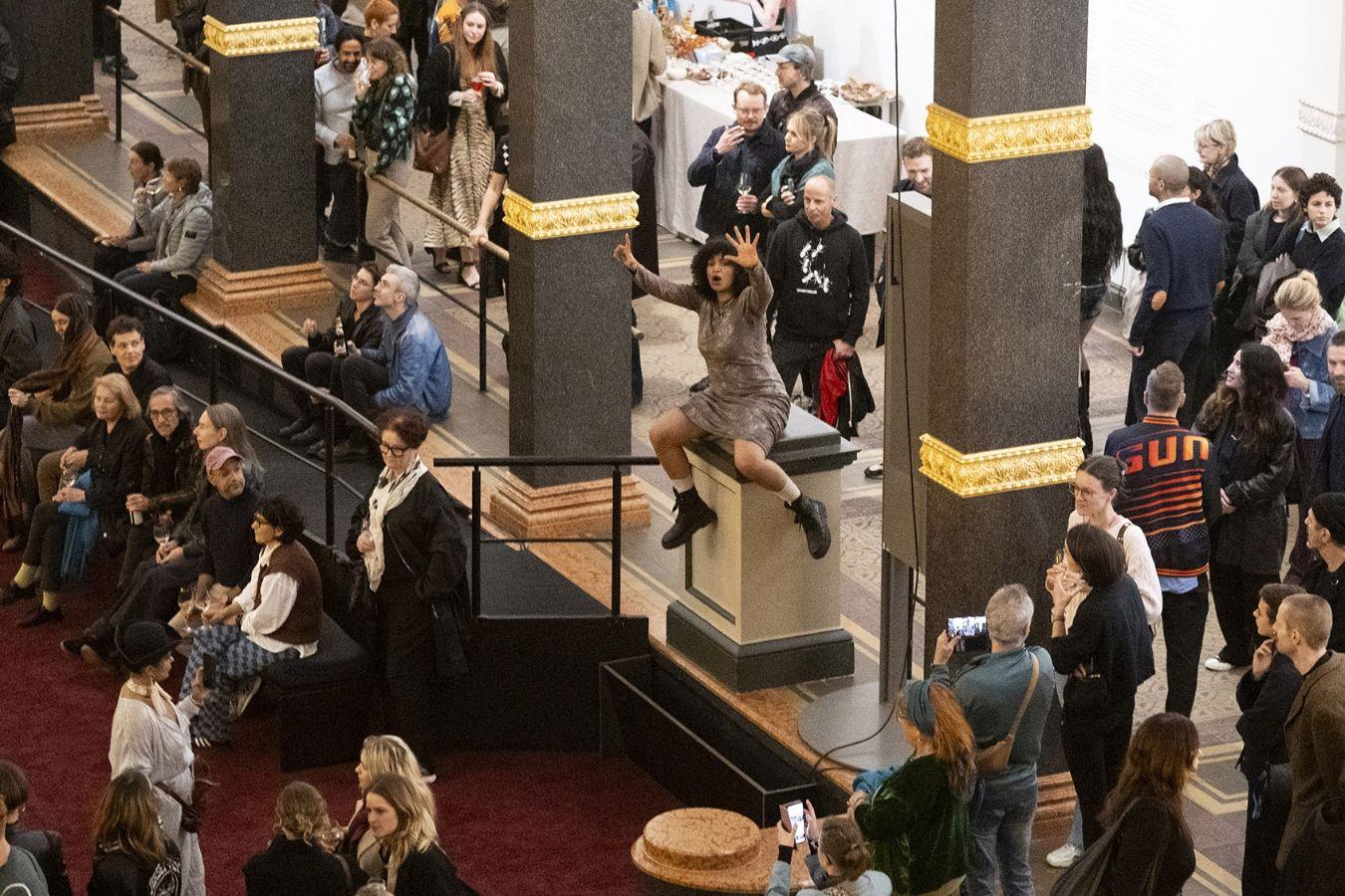 Der volle Lichthof des Gropius Bau, bei der Eröffnung der Ausstellung Ligia Lewis: Ligia Lewis: I’M NOT HERE FORRRRR… In der MItte der Besuchenden sitzt eine performenen Person auf einem Sockel.