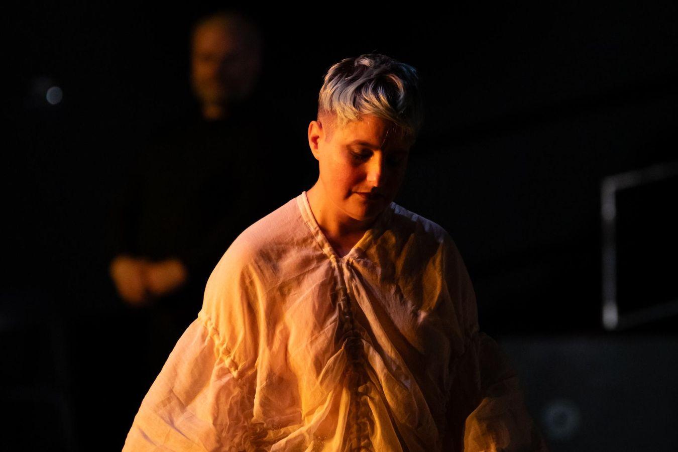 A person holding a walking stick in their right hand, dressed in a white, robe-like garment, illuminated from the left side by warm light.