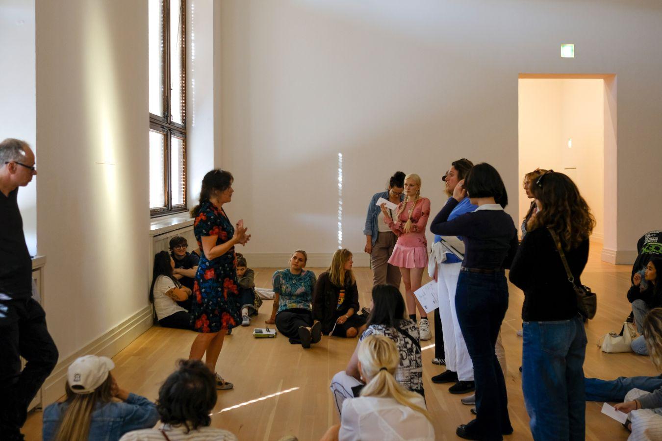 Several people sit and stand in an exhibition room and listen to a guided tour.