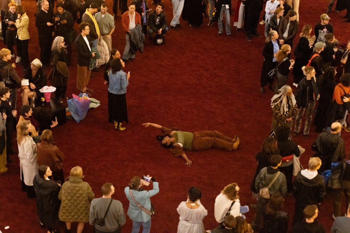 Der volle Lichthof des Gropius Bau, bei der Eröffnung der Ausstellung Ligia Lewis: Ligia Lewis: I’M NOT HERE FORRRRR… Eine Person performt auf einem roten Teppich, umgeben von Zuschauenden.