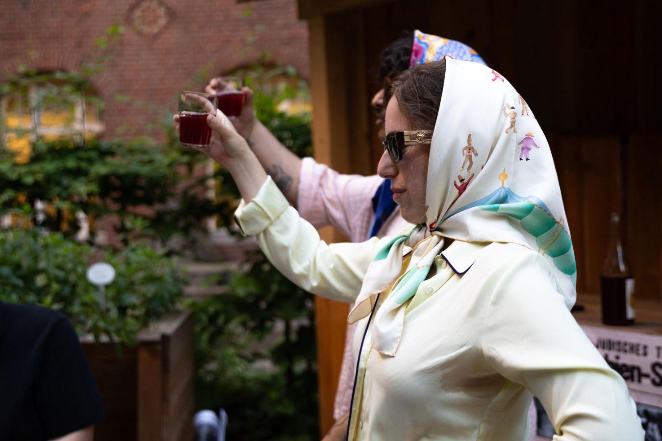 In profile, two figures raise their brimming glasses in a gesture of celebration, each adorned with a headscarf and shaded by dark glasses.