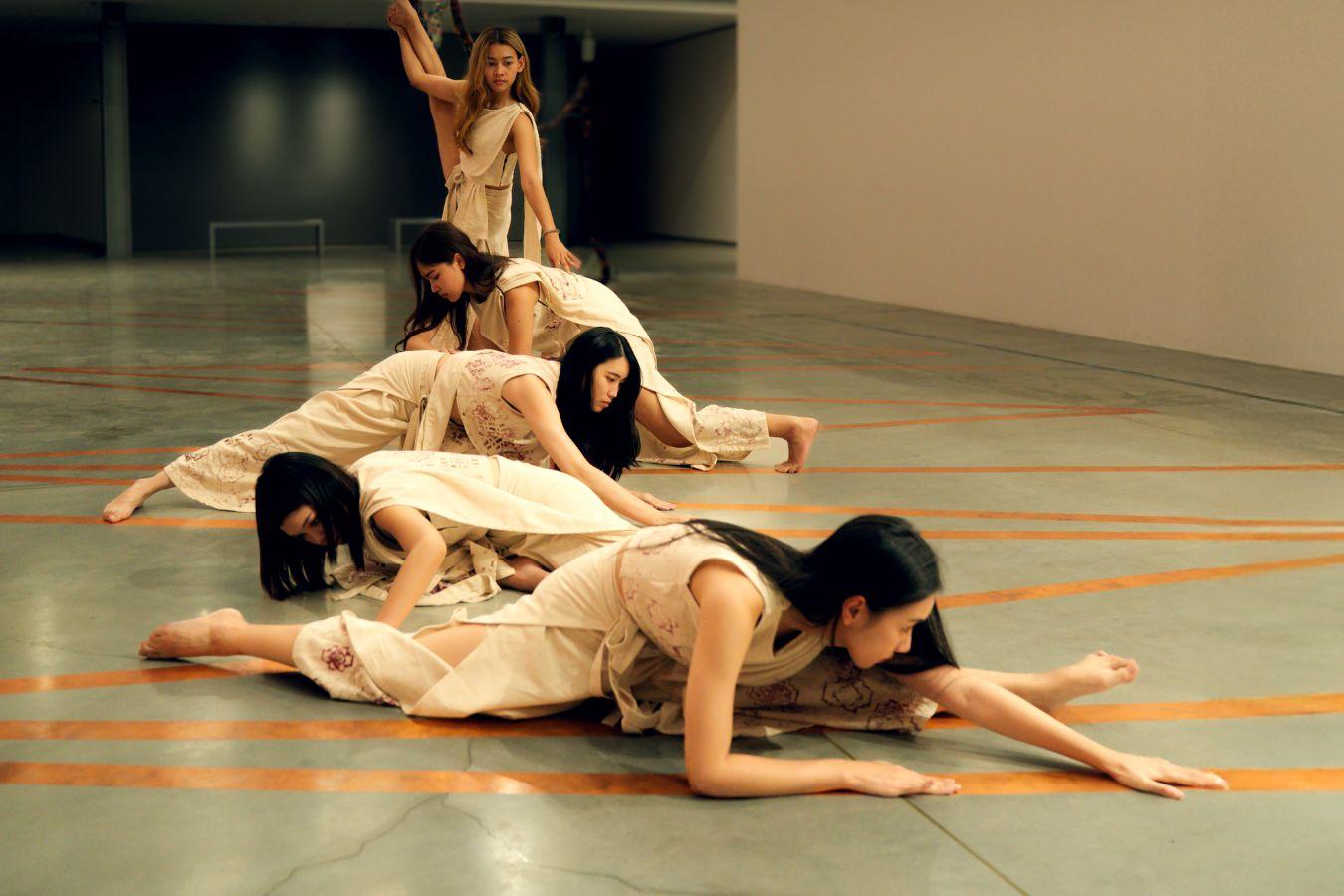 Fünf Performer*innen befinden sich in einer Halle und nehmen, in einer in die Tiefe weisenden Reihe, verschiedene tänzerische Haltungen ein.