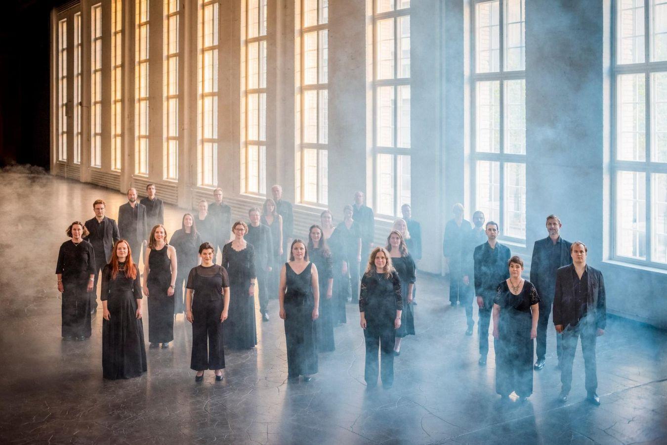 Der Helsinki Chamber Choir steht in einer großen Industriehalle. Durch die Fenster fällt gelb-bläuliches Licht, das sich im leichten Nebel fängt.