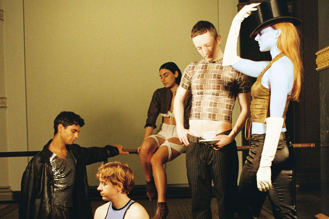 Five performers of Alex Baczyński-Jenkins "Malign Junction (Goodbye, Berlin)" stand and sit on a wooden platform. They look thoughtfully in different directions.