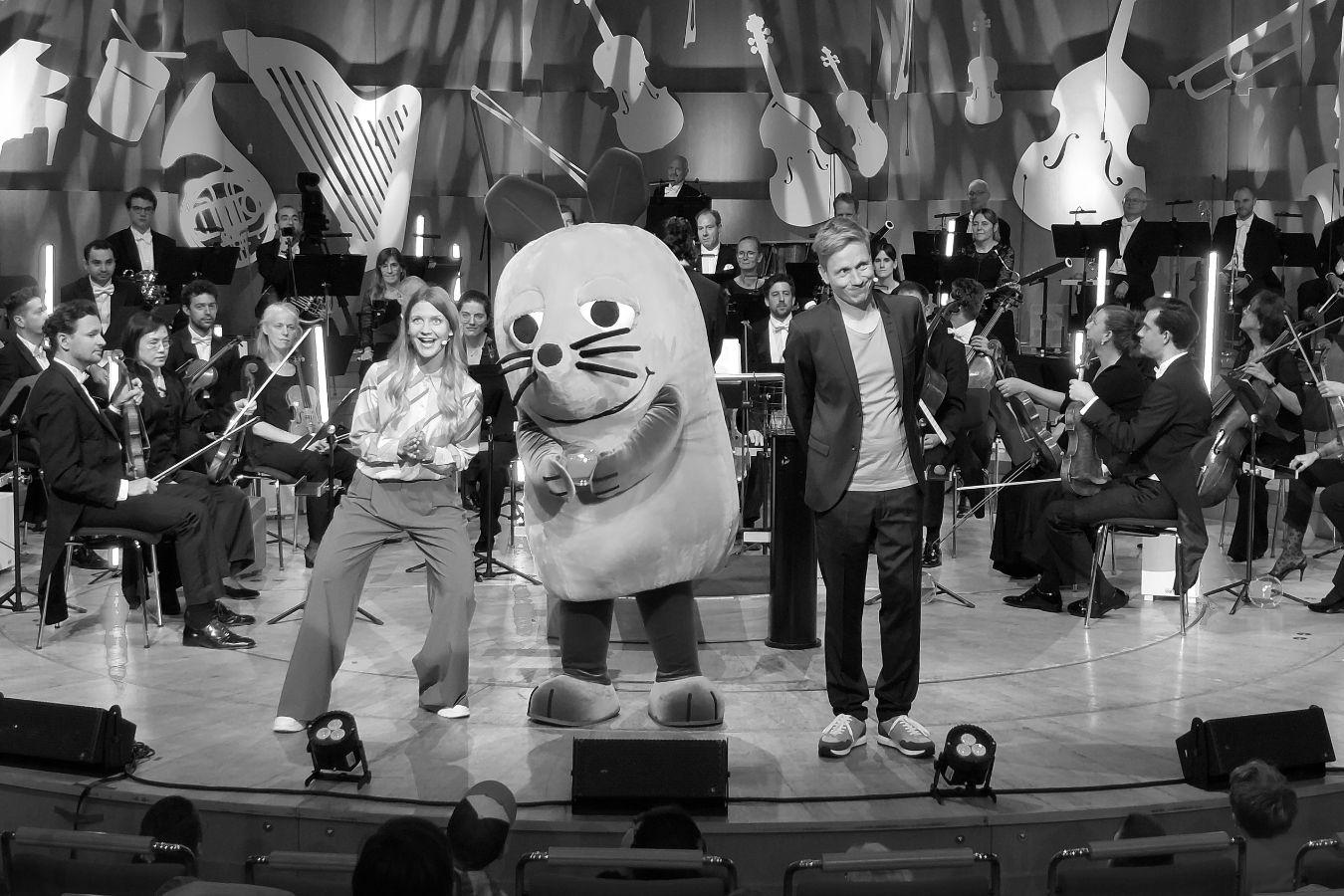 Zwei Moderator*innen, eine Maus und Orchester im Hintergrund auf einer Bühne