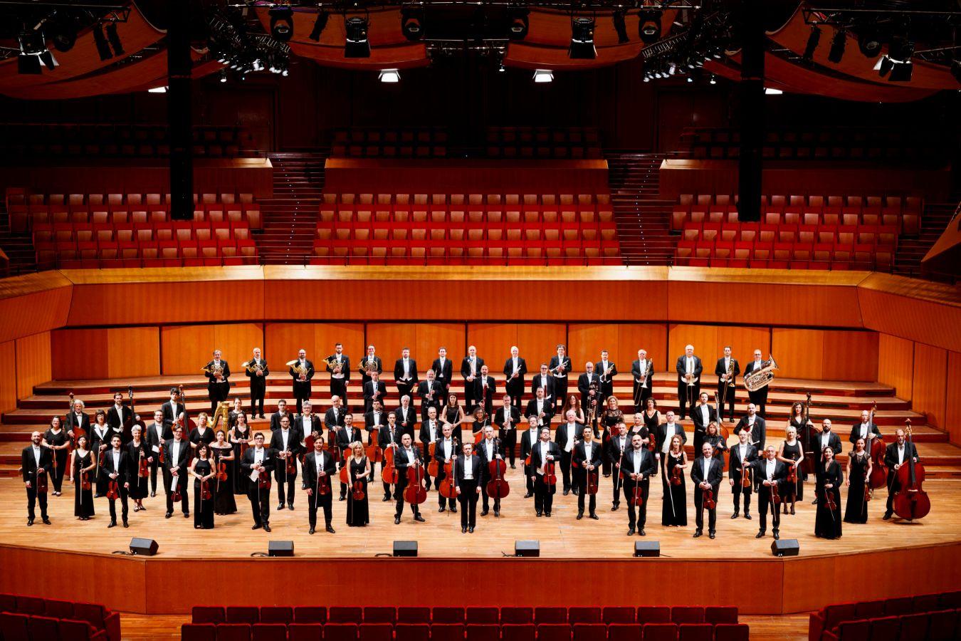 Musicians from a large orchestra stand with their instruments in a concert hall and look into the camera.