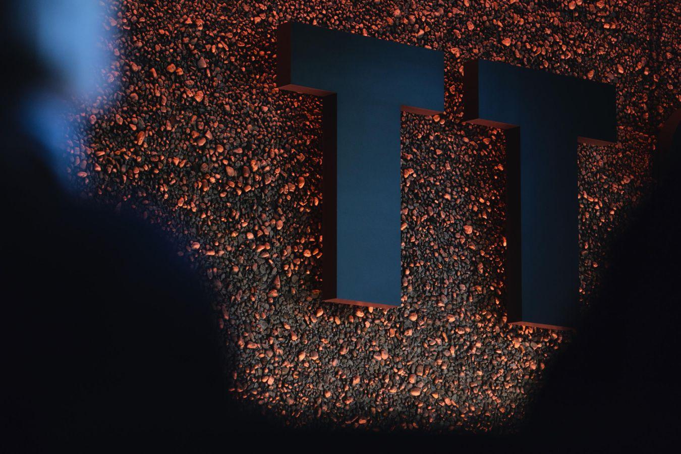 An einer orange beleuchteten Kieselwand hängen die Buchstaben "TT" in schwarzer Farbe.