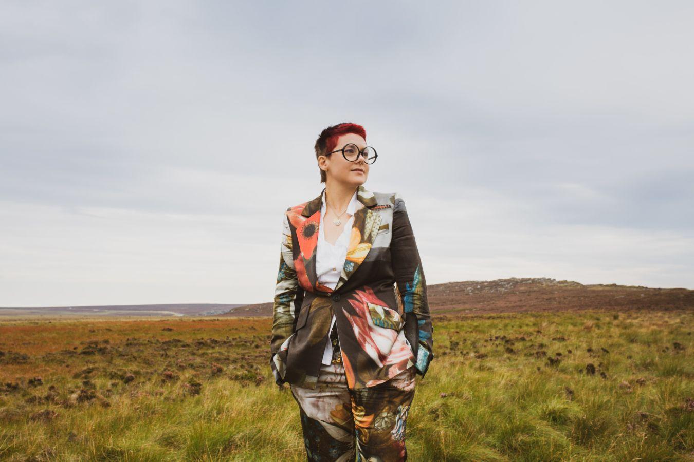 Bowler is standing in a meadow. She is wearing large glasses and a suit with large floral patterns.