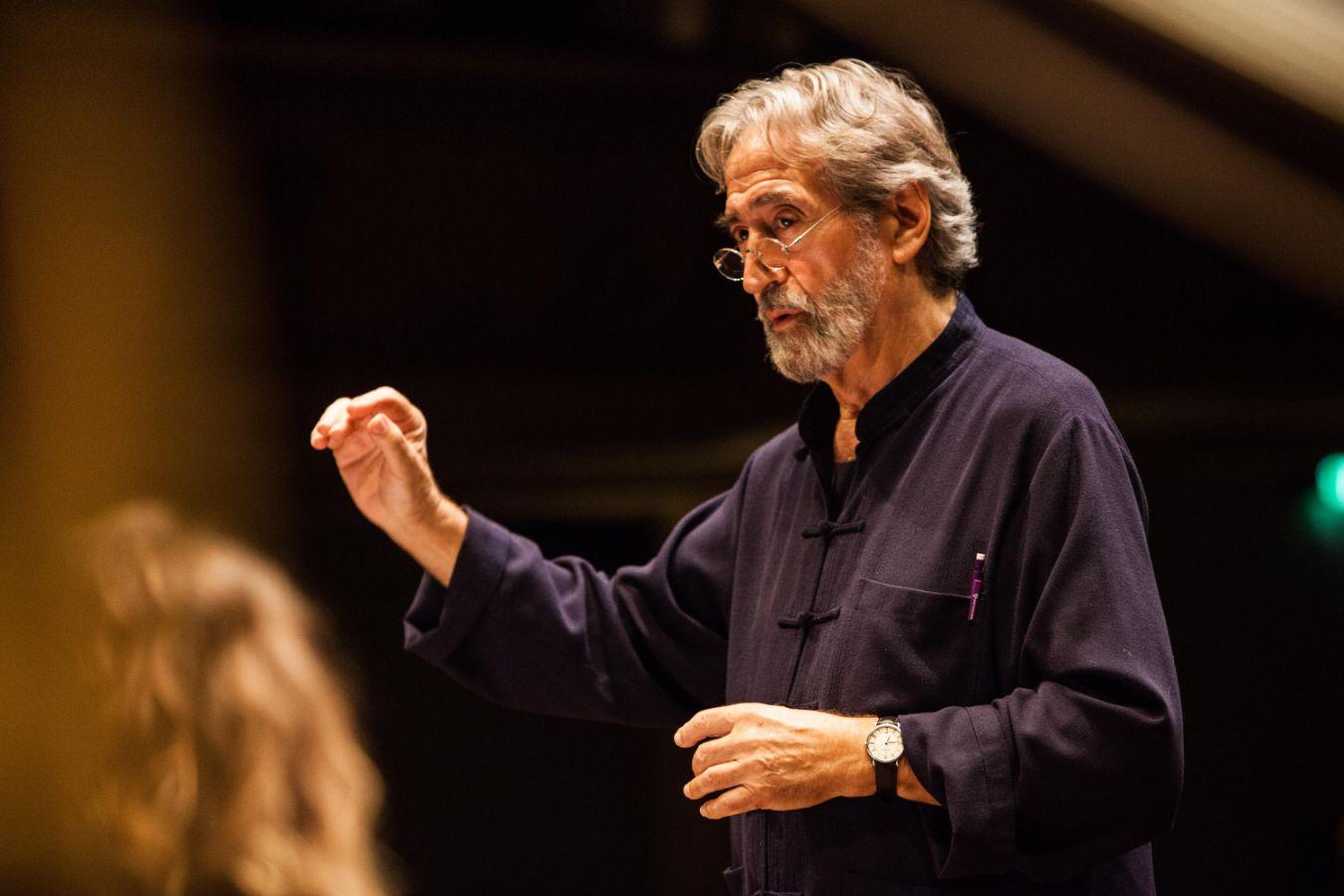 A man with grey hair and glasses is conducting, his right hand raised.