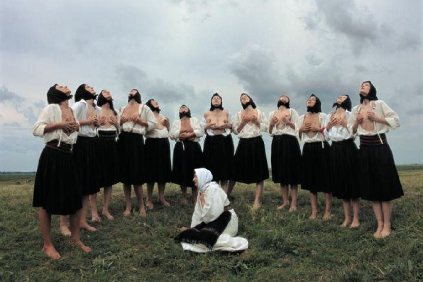 Frauen mit schwarzen Kopftüchern und knielangen Röcken massieren ihre nackten Brüste. Dabei schauen sie in den Himmel.