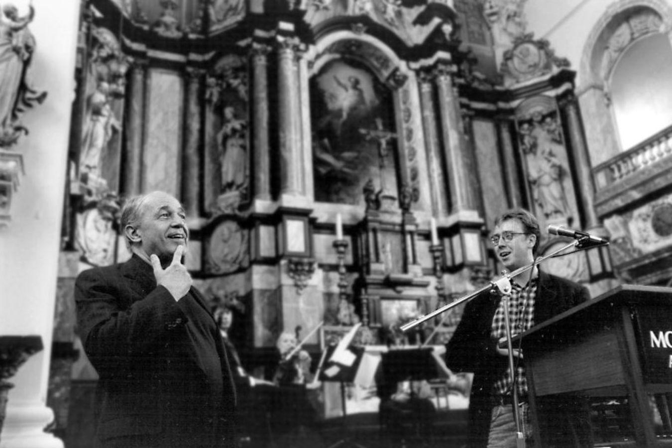An orchestra is playing in a church. Two men are standing in the foreground.