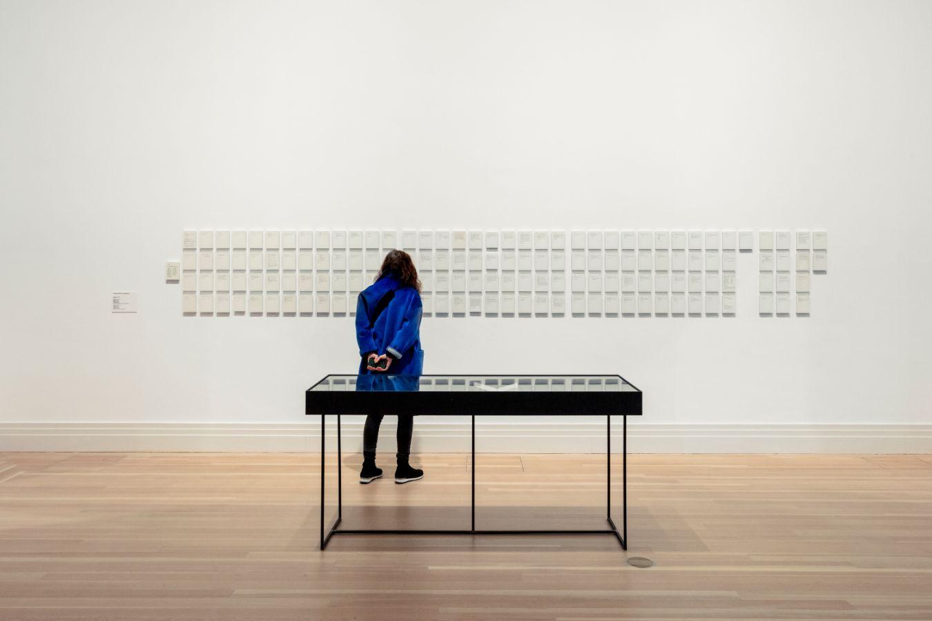 A person stands in an exhibition room in front of many small frames, in which small pieces of paper can be recognised.