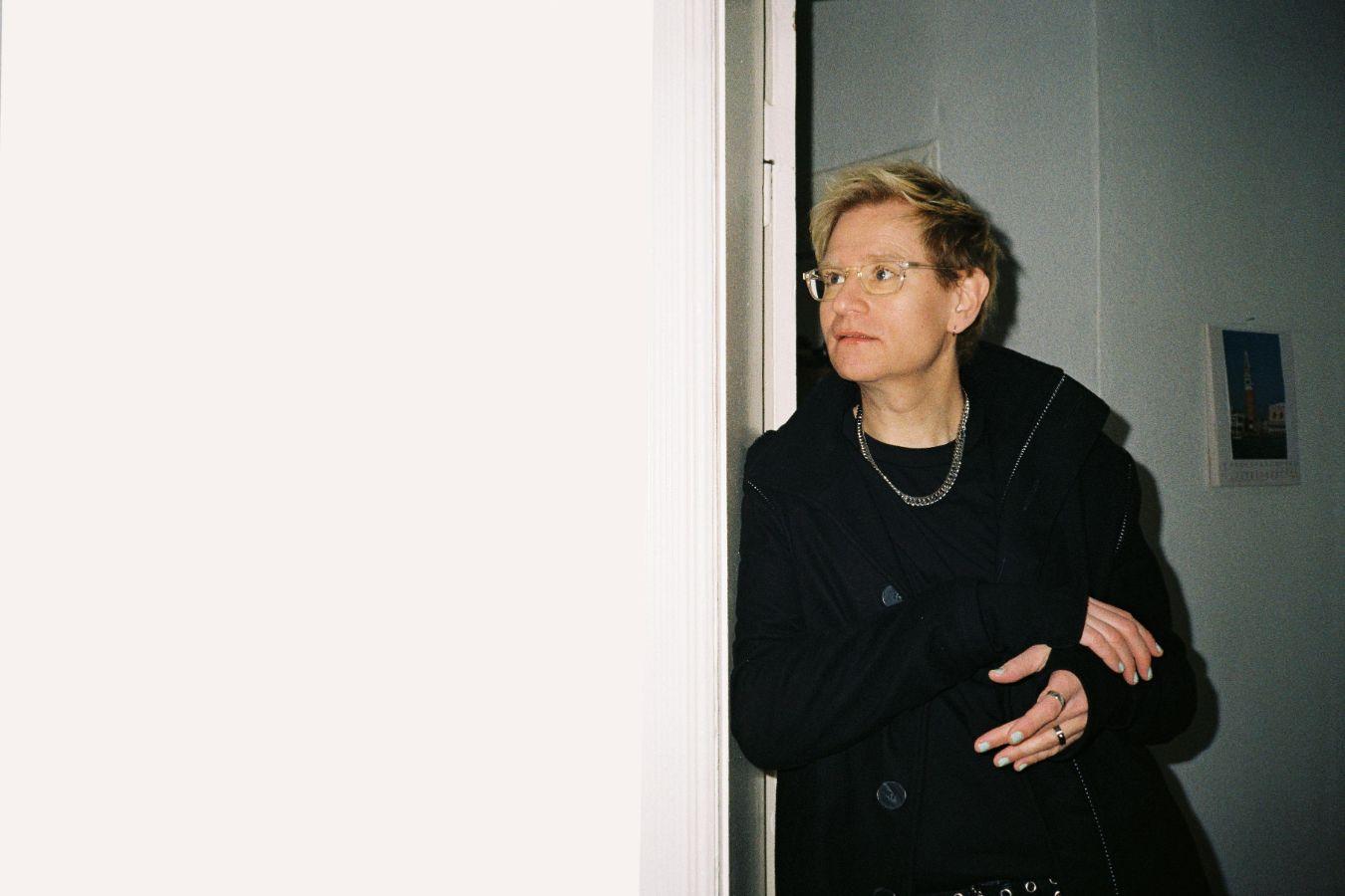 Luxa M. Schüttler is leaning against a door frame. Luxa's hair is short and dyed blonde, and their fingernails are painted. Luxa wears glasses.