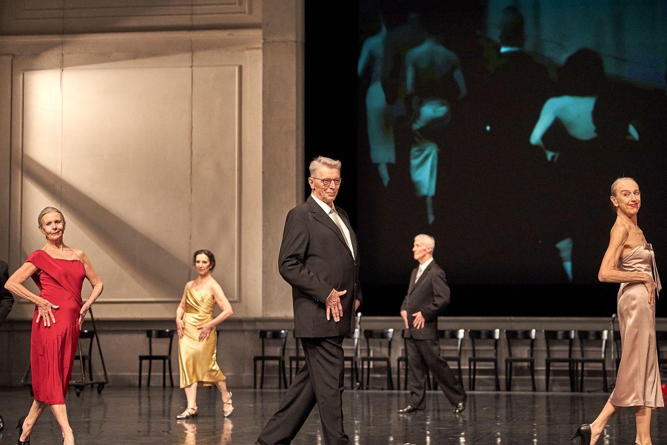 Seven people in formal wear dance in a circle in front of a video projection in black and white.