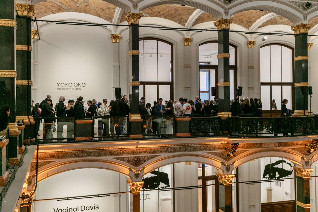Eine Menschenlange entlang des Obergangs im Lichthof des Gropius Bau. An der Wand steht der Ausstellungstitel „YOKO ONO: MUSIC OF THE MIND“.