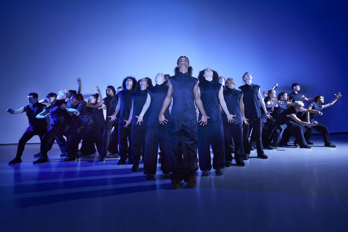 Several dancers stand formatted as a triangle in front of a blue background