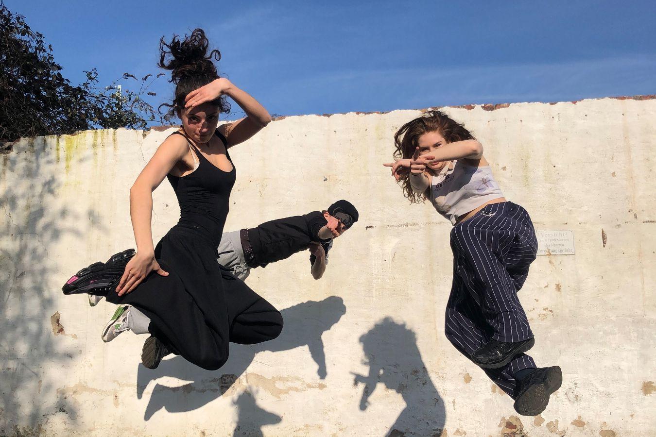 Drei Tänzer*innen springen vor einer weißen Mauer in die Luft.