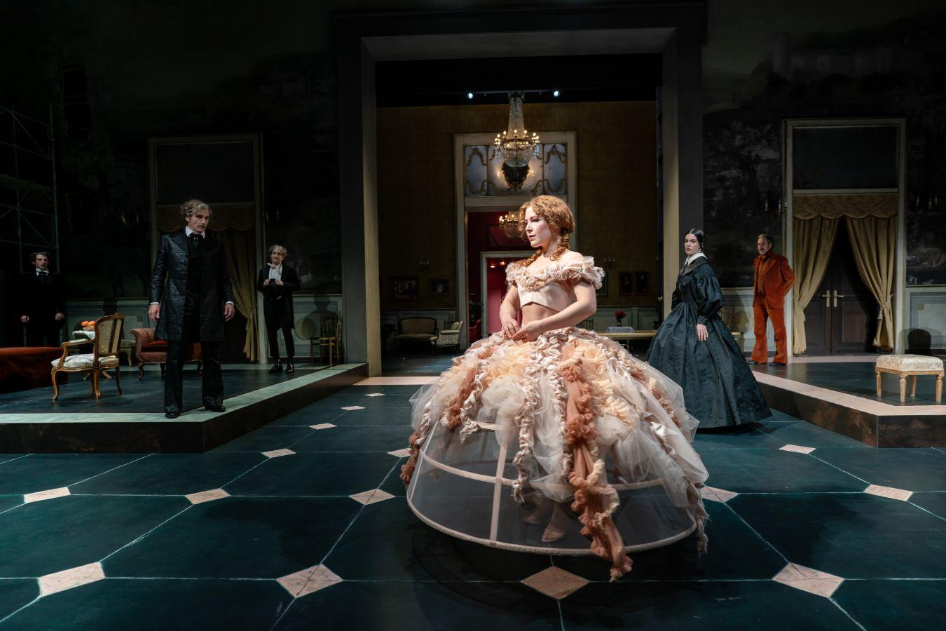 Eine junge Frau in einem Reifrock steht in einem Salon, im Hintergrund sind weitere Personen in historischen Kostümen