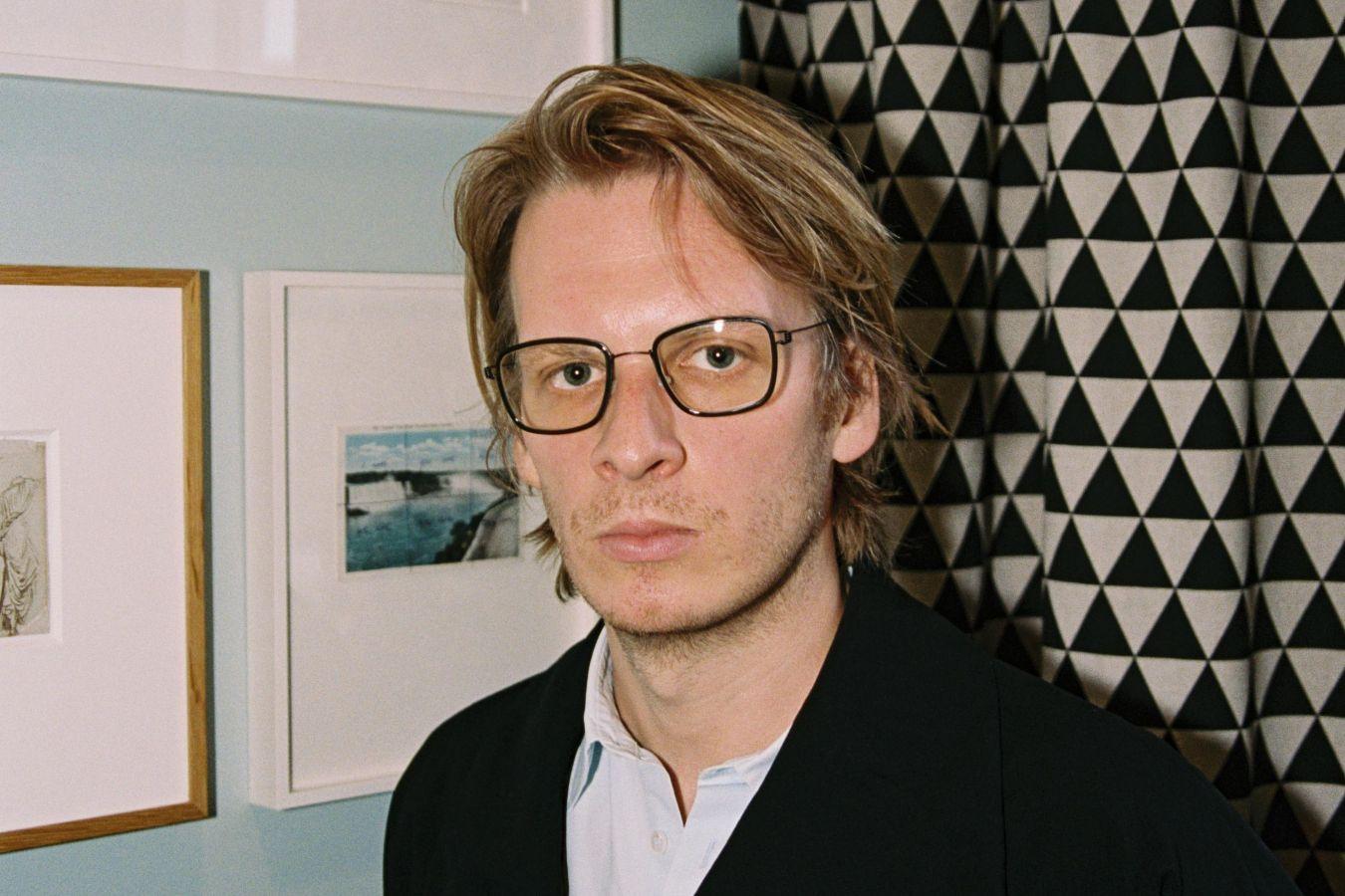 Portrait of a male‑presenting person wearing glasses, with a patterned curtain and picture frames in the background.