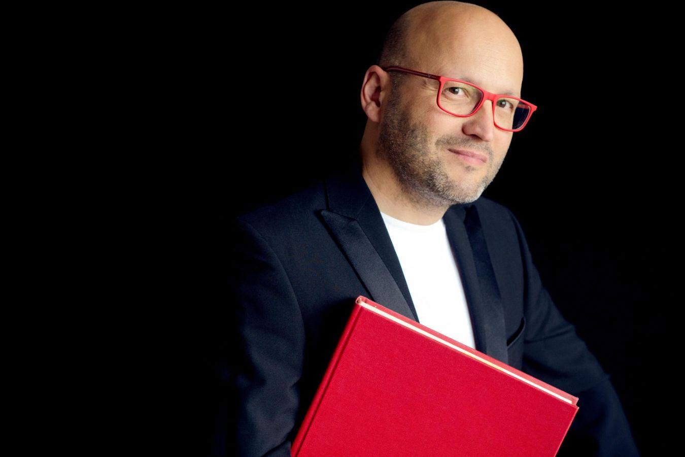 Ein Mann mit roter Brille und einem großen roten Buch unter dem Arm schaut in die Kamera.