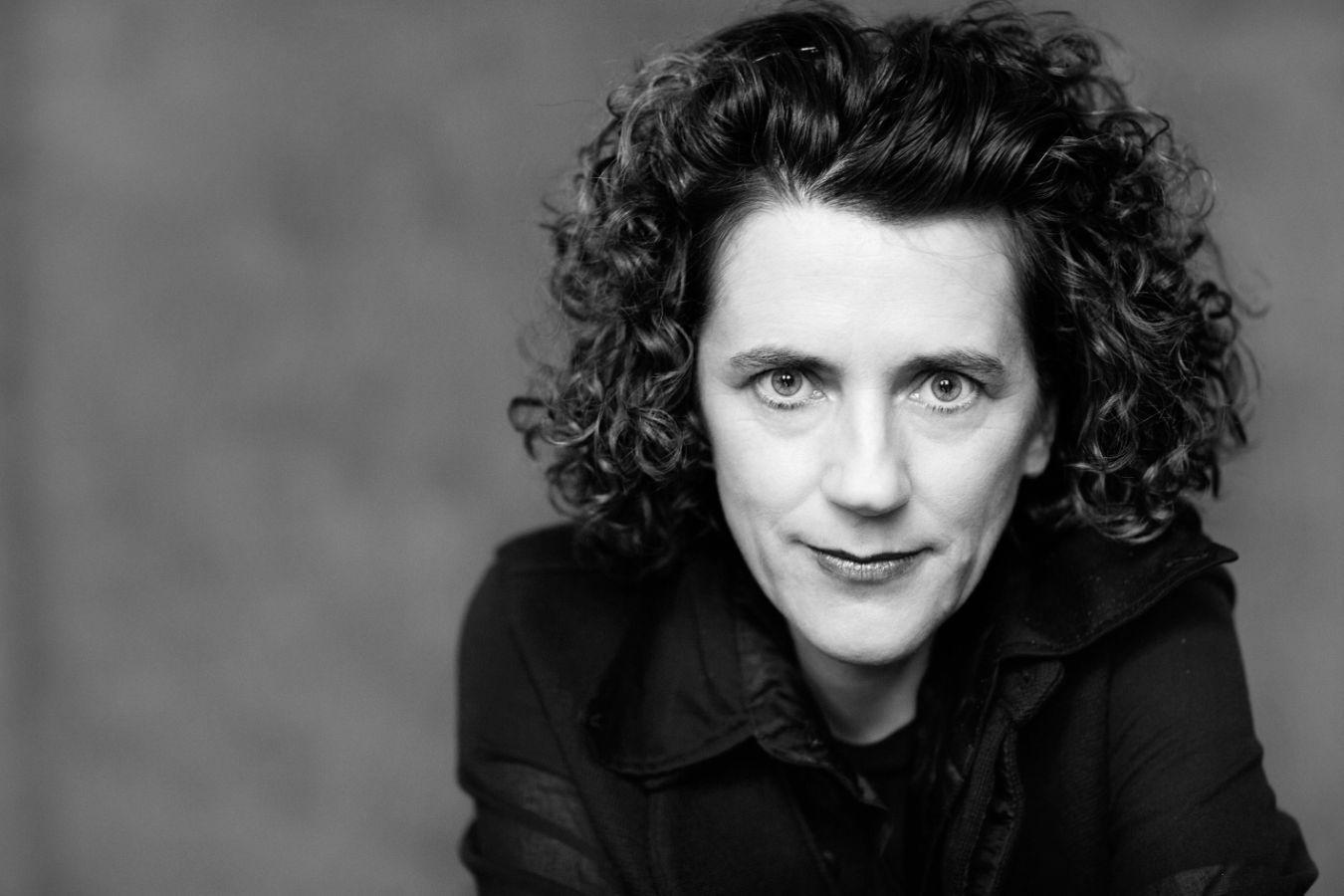 Eine Frau mit dunklen lockigen Haaren schaut sehr direkt mit einem Lächeln auf den Lippen in die Kamera.