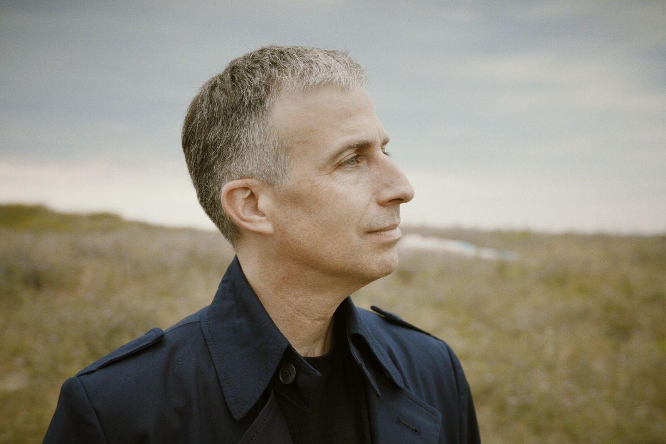 Das Profil eines Mannes mit kurzen Haaren ist zu sehen. Er steht in einer kargen Landschaft, der Himmel ist bewölkt.