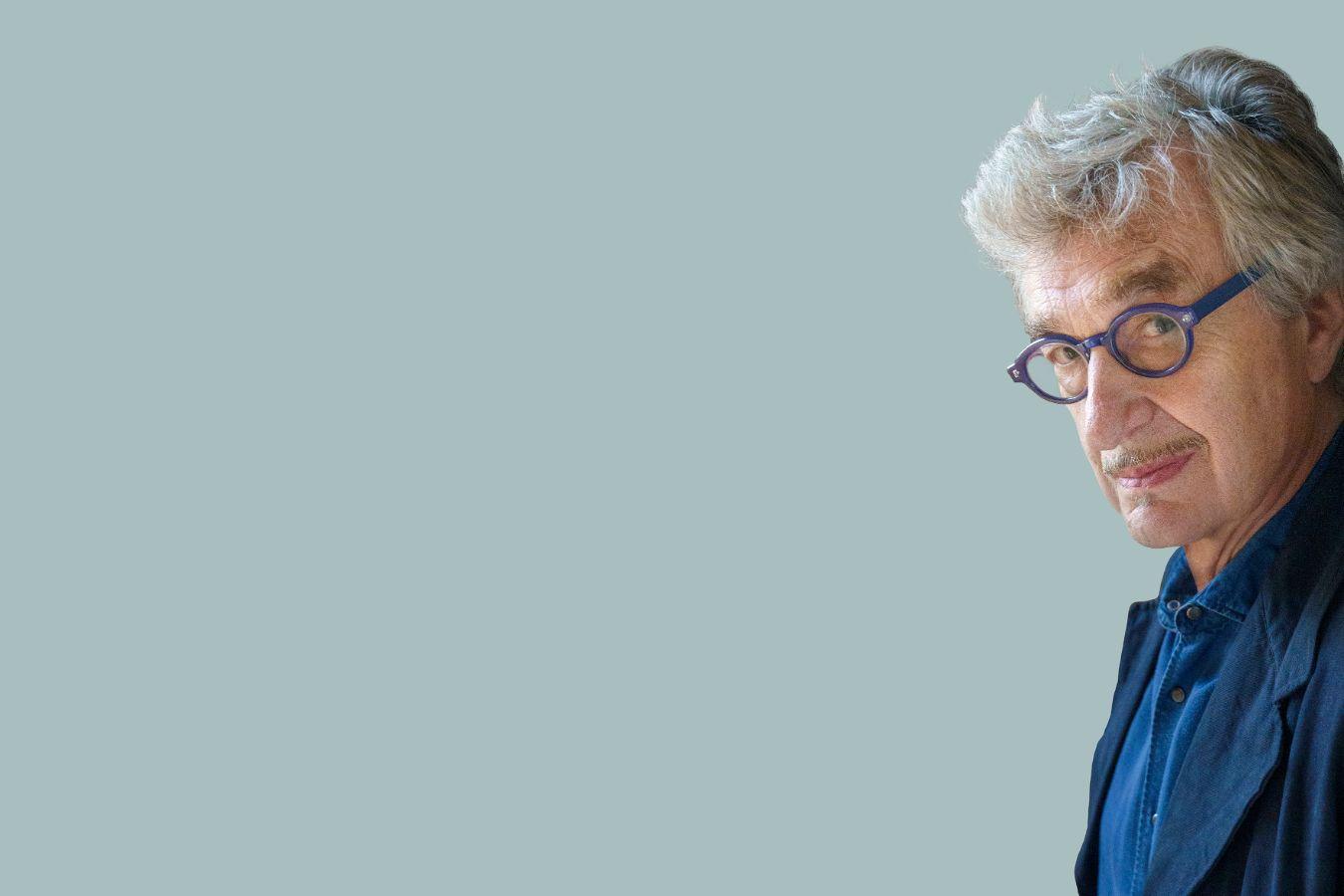 Ein Mann mit grauen Haaren und blauer Brille schaut schmunzelnd aus dem rechten Bildrand in die Kamera.