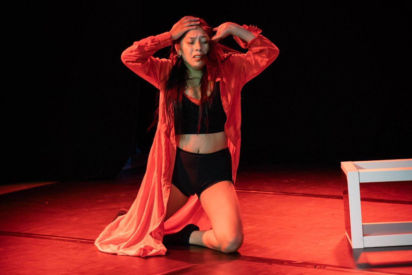 The dancer sits on her knees, her upper body upright, on the dark stage. Red light falls on her, she is wearing black clothes and a long red coat.