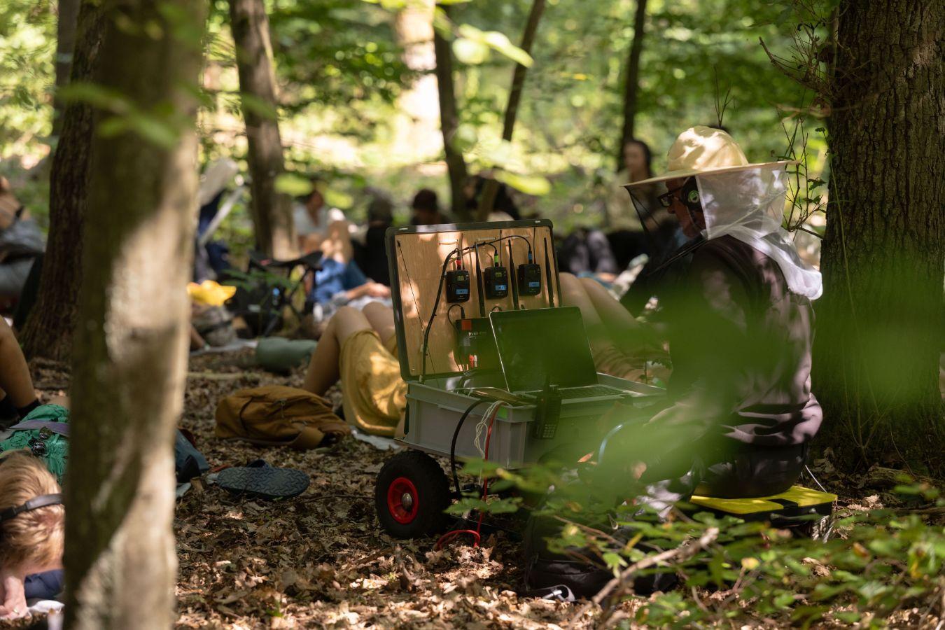 Audience lying and sitting on the forest floor, between them sits a man wearing a hat with a mosquito net